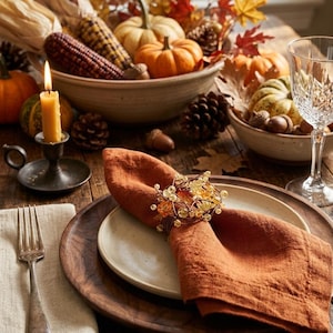 May include: A burnt orange linen napkin folded on a cream ceramic plate, resting on a dark wooden charger. The napkin is secured with a decorative wire ring featuring small amber-colored beads and metallic accents, creating a seasonal table setting.
