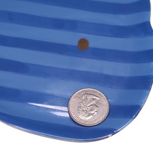 May include: A glossy blue ceramic dish featuring horizontal stripes in a darker shade of blue. A small brown circular mark is visible on the surface. A United States quarter coin is placed on the dish to provide a scale reference for its size.