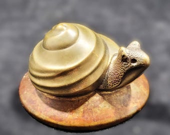 May include: A small, gold-colored snail sculpture with a detailed shell and a textured body. The snail is sitting on a round, brown base.