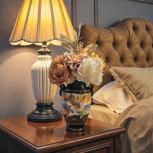 May include: A bedside table with a lamp and a vase of flowers. The lamp has a pleated shade and a cream-colored base with vertical lines. The vase is black with a floral design and holds a bouquet of peach and white flowers.