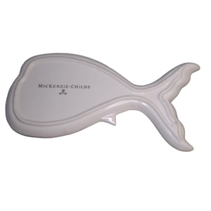 May include: A white ceramic dish shaped like a whale, featuring a smooth, glossy finish and a defined tail section. The text MACKENZIE-CHILDS is printed in black on the center of the dish, accompanied by a small crest logo below the brand name.