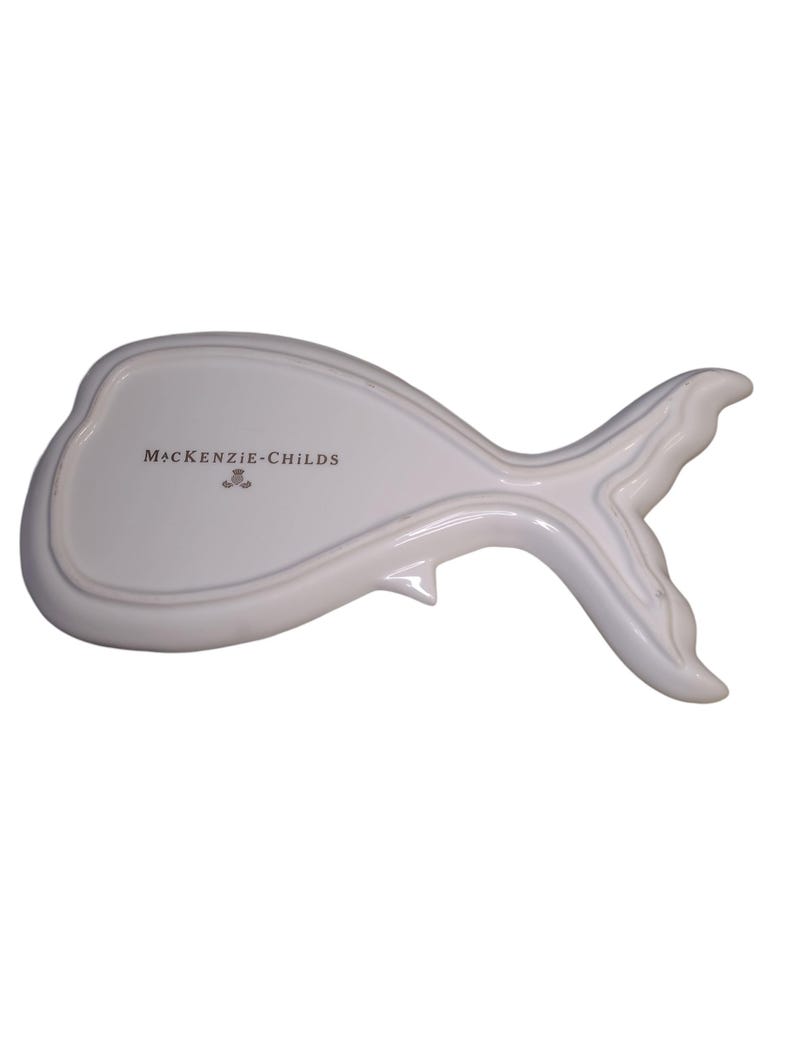 May include: A white ceramic dish shaped like a whale, featuring a smooth, glossy finish and a defined tail section. The text MACKENZIE-CHILDS is printed in black on the center of the dish, accompanied by a small crest logo below the brand name.