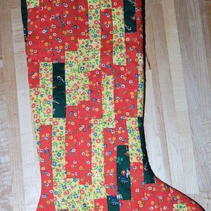 May include: A quilted Christmas stocking with a red floral pattern, accented with green and yellow stripes. The stocking features a loop for hanging and is decorated with festive imagery.