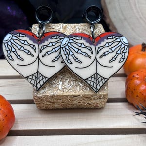 May include: A pair of black heart-shaped earrings with a skeleton hand design. The earrings are made of acrylic and have a black metal hook.