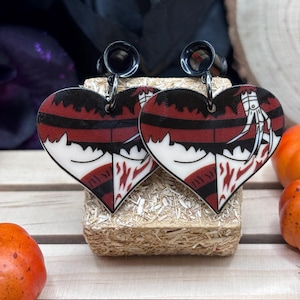 May include: A pair of black heart-shaped earrings with a bloody, red and white design. The earrings feature a black metal plug and a dangling heart charm.