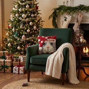 May include: A dark green velvet armchair featuring a square throw pillow with a cat face and the text MEOW, topped with a red and white polka dot Santa hat. A cream-colored knit throw blanket is draped over the arm of the chair.