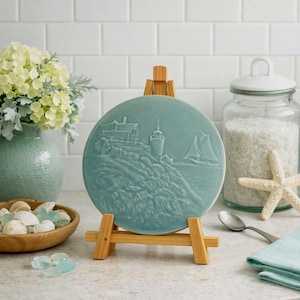 May include: A round, teal-colored ceramic plate with a raised relief design of a coastal scene, including a lighthouse, house, and sailboat. The plate is displayed on a small wooden easel. Other coastal decor includes shells, a starfish, and a vase of flowers.