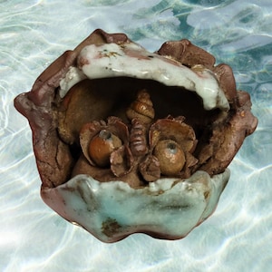 May include: A small, ceramic sculpture of a creature with a shell-like head and a mouth full of small, brown, round objects. The sculpture is glazed in shades of brown, white, and blue.