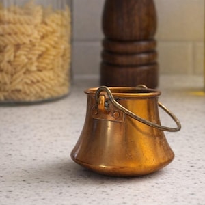 May include: A small, antique copper pot with a curved handle. The pot has a rounded base and a flared rim. The handle is attached to the pot with small rivets. The pot is sitting on a speckled countertop.