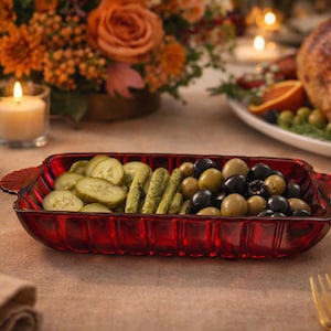 May include: A red glass serving dish filled with sliced pickles, whole pickles, and olives. The dish is on a table set for a meal, with a turkey, flowers, and candles in the background.