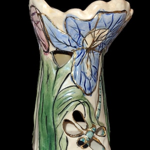 May include: Hand-painted ceramic vase featuring a blue iris flower design with gold accents. The vase has a dragonfly cutout and green leaves. The artist's signature, "Heather Goldmine", is visible on the base.