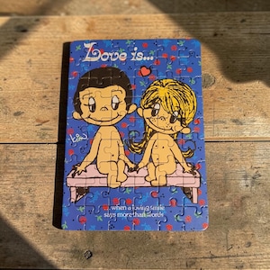 May include: A rectangular jigsaw puzzle featuring the Love is... cartoon characters sitting on a bench. The blue background has a red floral pattern. Text reads Love is... and when a loving smile says more than words. The puzzle is assembled.