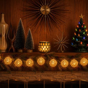 May include: A string of round, woven metal lanterns with warm internal lights draped across a mantelpiece. The display includes a ceramic Christmas tree with colorful bulbs, two bottle brush trees, a candle holder, and a starburst clock.