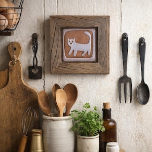 May include: A square wooden frame containing a ceramic tile with a white cat illustration on a brown background. The frame is mounted on a white wall alongside various kitchen utensils, including wooden spoons, a whisk, and metal tools.