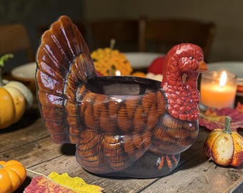 Op de afbeelding: Een keramische kalkoen-vormige serveerschaal met bruine en rode tinten. De schaal heeft een gedetailleerd veermotief en een hol midden. Het staat op een houten tafel met pompoenen en herfstbladeren, wat een Thanksgiving-thema oproept.