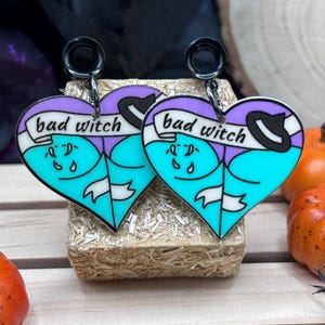 May include: A pair of black heart-shaped earrings with a turquoise and purple design. The earrings feature a black witch's hat and the text "bad witch" on a white banner.