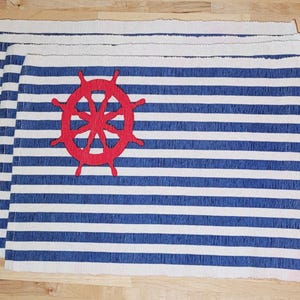 May include: Set of four nautical-themed placemats. Each placemat features blue and white horizontal stripes and a red ship's wheel emblem. The placemats are rectangular and made of a woven material.