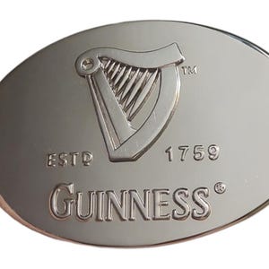 May include: Oval-shaped, silver-colored metal emblem featuring the Guinness logo. The logo includes a harp, the word "Guinness", and the year "1759". The emblem has a polished, reflective surface.