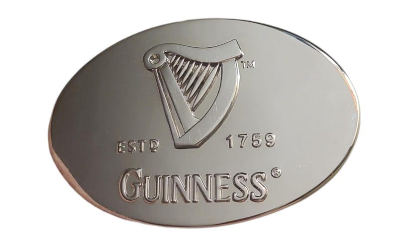 May include: Oval-shaped, silver-colored metal emblem featuring the Guinness logo. The logo includes a harp, the word "Guinness", and the year "1759". The emblem has a polished, reflective surface.
