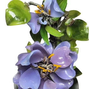 May include: A glass flower arrangement featuring a cluster of lavender-colored blooms with yellow stamens and green leaves. The flowers are in a small, gray ceramic pot, creating a decorative accent.