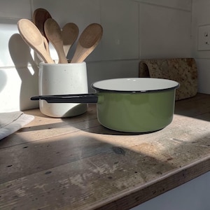 May include: A light green enamel saucepan with a black handle sits on a wooden surface. A white speckled ceramic utensil holder with wooden spoons is to the left. A cork trivet is in the background.