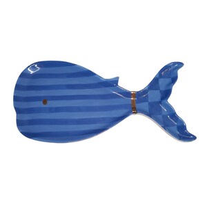 May include: A whale-shaped ceramic dish featuring a blue and light blue horizontal striped pattern on the body and a blue checkered pattern on the tail. A thin copper-colored band separates the body from the tail, and a small brown dot marks the eye.