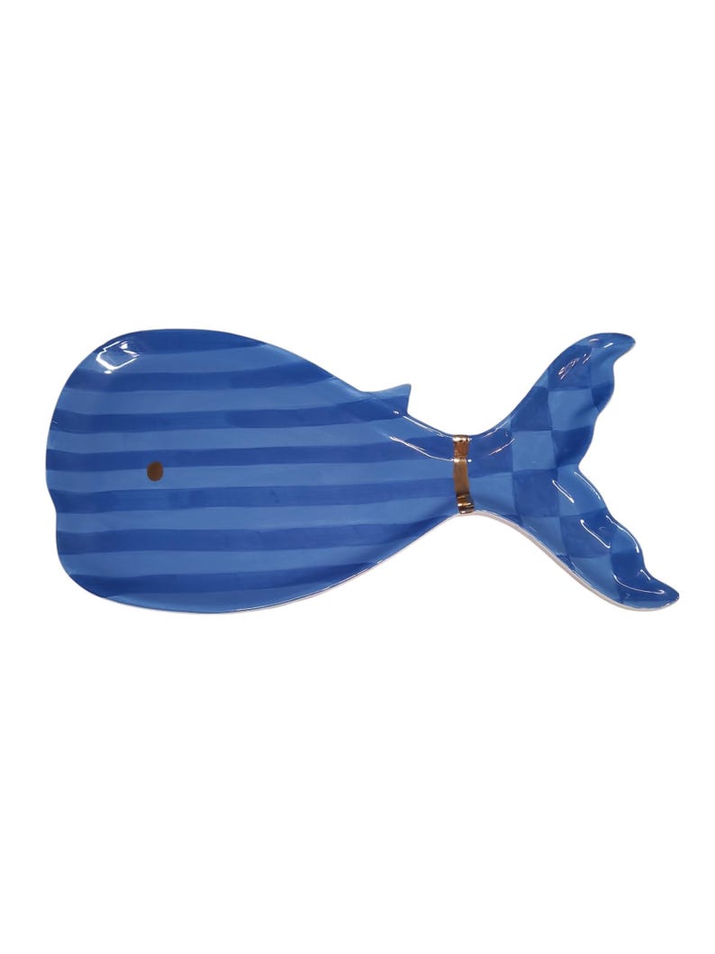 May include: A whale-shaped ceramic dish featuring a blue and light blue horizontal striped pattern on the body and a blue checkered pattern on the tail. A thin copper-colored band separates the body from the tail, and a small brown dot marks the eye.
