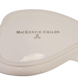 May include: An off-white ceramic dish with a smooth, glossy finish. The center features the text MACKENZIE-CHILDS printed in a dark gray font, accompanied by a small, detailed thistle icon positioned directly below the brand name.
