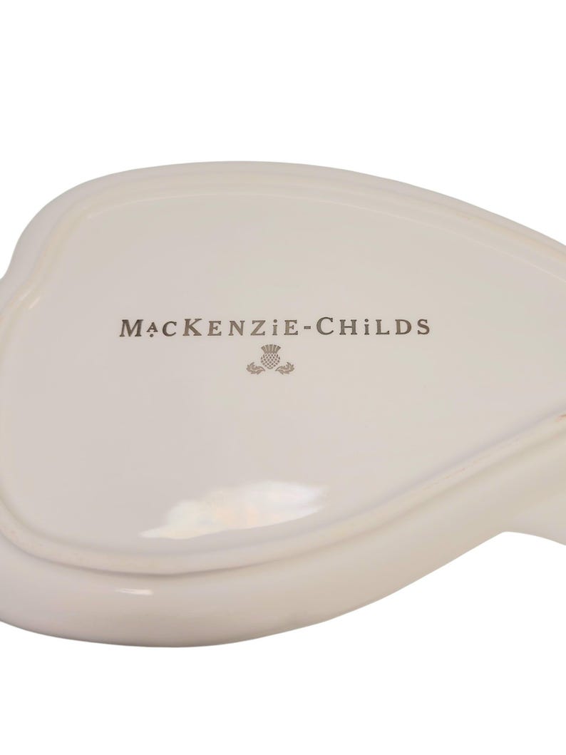 May include: An off-white ceramic dish with a smooth, glossy finish. The center features the text MACKENZIE-CHILDS printed in a dark gray font, accompanied by a small, detailed thistle icon positioned directly below the brand name.