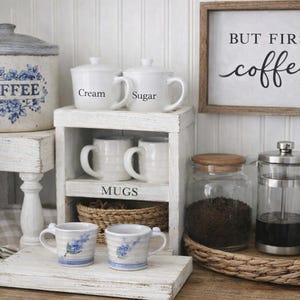 May include: A rustic coffee station with a sign that reads "BUT FIRST coffee". It features a large coffee canister, white mugs, a French press, and a jar of coffee beans. The mugs are labeled "Cream" and "Sugar".