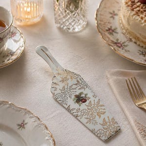 May include: A white and gold floral patterned cake server with a decorative handle. The server is placed on a white tablecloth, alongside a plate, a teacup, and a cake.
