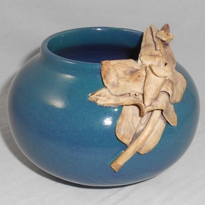 May include: A teal blue ceramic vase with a decorative flower design made of light brown clay.