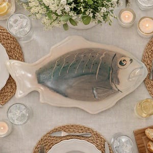 May include: A ceramic serving platter shaped like a fish with a cream-colored border and a central body featuring blended shades of blue, grey, and brown. The fish design includes a painted eye, fins, and scale details on a glossy, smooth surface.