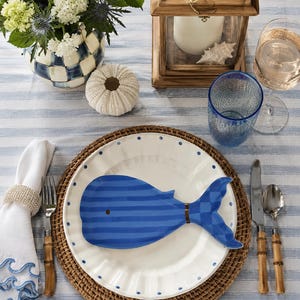 May include: A white ceramic dinner plate with blue polka dots, topped with a blue striped whale-shaped dish. The setting includes a woven charger, a white napkin with blue wave embroidery, bamboo-handled silverware, and a blue textured glass.