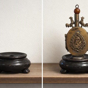 May include: A brass oval-shaped container with a central teardrop-shaped relief featuring a bird, flanked by two rooster figures at the top and a brown bead finial, resting on a dark brown, three-legged circular wooden base with a central hole.
