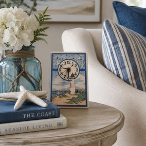 May include: A rectangular tabletop clock featuring a lighthouse illustration with a sandy shore, blue sky, and flying seagulls. The clock face has black numerals and dark hands. The design is framed by a blue border on a light-colored background.