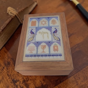 May include: Wooden box with a decorative tile top featuring religious symbols, including a Star of David, a menorah, and peacocks. The box has a light brown wooden frame and a detailed, colorful design.