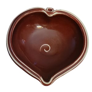 May include: A heart-shaped ceramic bowl in a rich, reddish-brown hue. The bowl features a white spiral design in the center and a white trim along the rim. It has a small spout and a loop on the side, suggesting it may be a serving dish.