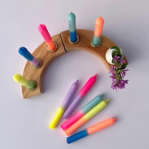 Könnte beinhalten: Ein hölzerner Kerzenhalter in Halbkreisform, der sieben farbige Kerzen hält. Die Kerzen sind eine Mischung aus leuchtenden Farben, darunter Rosa, Orange, Blau und Gelb. Eine kleine weiße Vase mit lila Blumen ist ebenfalls vorhanden.