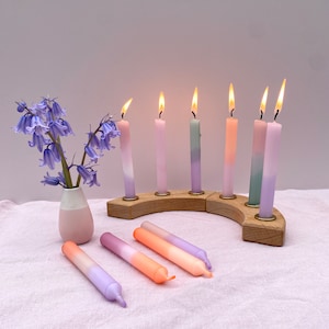 Könnte beinhalten: Ein Holz-Menora mit sieben brennenden Kerzen in Violett-, Rosa- und Grüntönen. Die Kerzen sind in einem Halbkreis auf dem Menora angeordnet. Es gibt auch fünf kleinere Kerzen in Violett- und Rosatönen auf einer weißen Oberfläche.