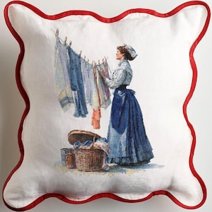 May include: Cushion featuring a vintage illustration of a woman hanging washing on a line. The pillow has a white background and a red, scalloped border.