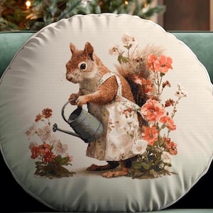 Puede incluir: Cojín decorativo redondo con una ilustración de una ardilla con delantal regando flores con una regadera de metal. El cojín tiene un borde verde claro y un diseño floral.