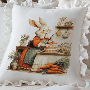 May include: White decorative pillow with a ruffled edge. The pillow features a cross-stitch design of a smiling rabbit in an orange and blue outfit, decorating a cake. Carrots and kitchen items are also visible in the illustration.