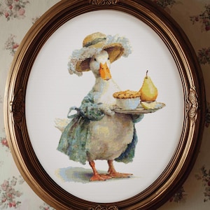 Puede incluir: Obra de arte enmarcada ovalada con un pato vestido con un gorro y un vestido, sosteniendo una bandeja con un pastel, una pera y una rodaja de limón. El pato está en un marco dorado sobre un fondo de papel pintado floral.