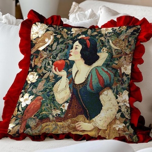 Puede incluir: Cojín decorativo con un diseño de Blancanieves. El cojín tiene un fondo verde oscuro y floral con un borde rojo. Blancanieves se representa sosteniendo una manzana roja. El cojín está sobre una superficie blanca.