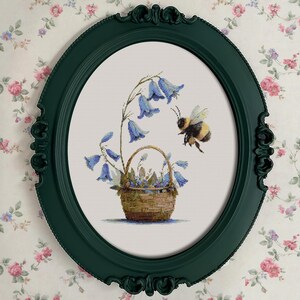 May include: A framed cross-stitch artwork featuring a basket of bluebell flowers and a hovering bee. The artwork is set within a dark green oval frame, against a floral wallpaper background. The bee is black and yellow.