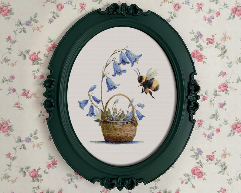 May include: A framed cross-stitch artwork featuring a basket of bluebell flowers and a hovering bee. The artwork is set within a dark green oval frame, against a floral wallpaper background. The bee is black and yellow.