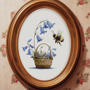 May include: An oval, gold-framed artwork featuring a cross-stitch design. The image depicts a basket of bluebell flowers with a bee hovering nearby. The background is a light cream color.