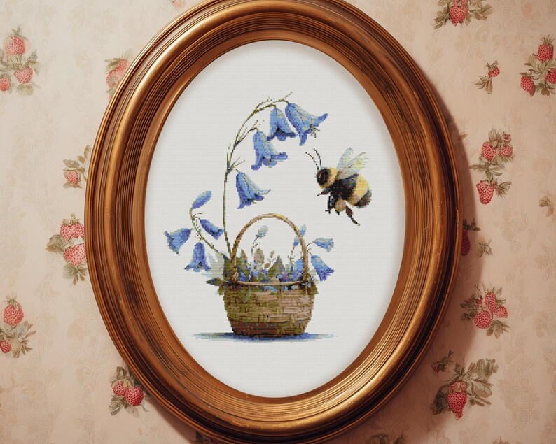 May include: An oval, gold-framed artwork featuring a cross-stitch design. The image depicts a basket of bluebell flowers with a bee hovering nearby. The background is a light cream color.