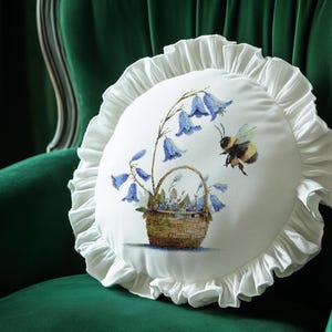 May include: A round white pillow with a ruffled edge, featuring a floral design of bluebell flowers and a bee. The flowers are in a woven basket. The pillow is set against a green velvet chair.